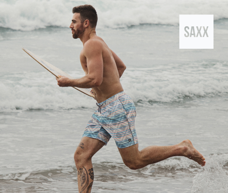 Man running on the beach with his surfboard, wearing geometric‑print swim shorts from SAXX.